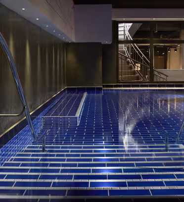 Modern inomhuspool med mörkblått kakel och krom ledstänger. Poolområdet är svagt upplyst, vilket skapar en reflekterande effekt på vattnet. Stora fönster i bakgrunden avslöjar en trappa och ytterligare utrymme inomhus.