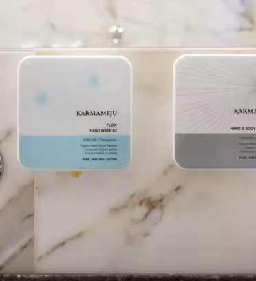 Två fyrkantiga Karmameju-automater på en marmorvägg; den vänstra innehåller "Flow" handtvätt i ljusblått, den högra "Chill" hand och bodylotion i grått. Båda är ovanför silverkranar märkta med ett "C" för kyla.