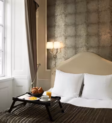 Ett mysigt hotellrum har en snyggt bäddad säng med vita kuddar och en beige sänggavel. En bricka med frukostartiklar, inklusive croissanter och juice, ligger på sängen. Två vägglampetter lyser upp utrymmet nära ett fönster med fördragna gardiner.