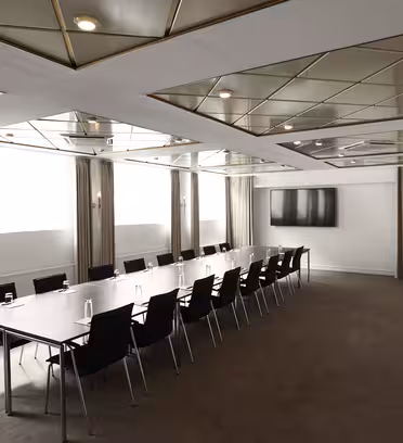 Ett modernt konferensrum med ett långt rektangulärt bord omgivet av svarta stolar. Taket har geometriska spegelpaneler och infälld belysning. En stor väggmonterad tv finns i rummets ände och gardiner täcker fönstren på sidoväggen.