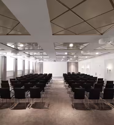 Ett modernt konferensrum med rader av svarta stolar mot en skärm framtill. Rummet är väl upplyst, med en geometrisk takdesign och beige matta. Det finns stora fönster längs ena sidan, täckta med ljusa gardiner.