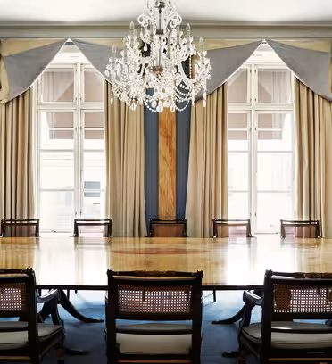 Elegant konferensrum med ett stort träbord omgivet av stolar. Dekorativa draperier och en kristallkrona framhäver utrymmet. Höga fönster med skira gardiner släpper in naturligt ljus i rummet, vilket förstärker den sofistikerade atmosfären.