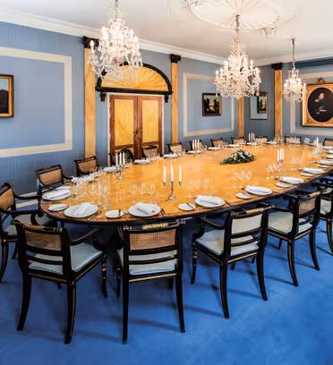 Elegant matsal med ett långt ovalt bord för en formell middag. Det blåväggiga rummet har ljuskronor, inramade målningar, speglar och snyggt arrangerade stolar. Bordet är prydt med tallrikar, glas och ett centralt blomsterarrangemang.