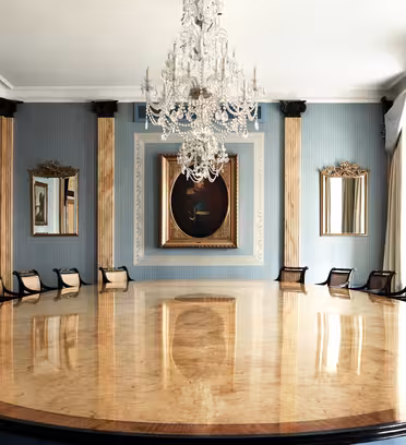 Elegant konferensrum med ett stort, polerat ovalt bord omgivet av svarta stolar. Rummet har ljusblå väggar, guldkantade speglar och en stor ljuskrona. Ett inramat porträtt hänger på den bortre väggen mellan höga fönster med draperier.