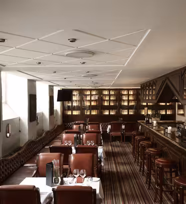 En stilren restauranginredning med röda läderstolar, en lång bar till höger och en rad bord med vita dukar till vänster. Hyllor med böcker och prydnadsföremål kantar bakväggen och mjuk belysning skapar en mysig atmosfär.