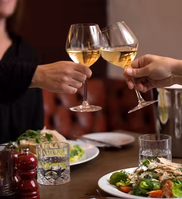 Två personer skålar med vita vinglas över ett dukat bord med salladsrätter och ett tänt ljus. En vinkyl finns i bakgrunden och miljön verkar vara en mysig restaurang.