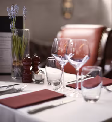 Elegant dukning i en restaurang med röda vadderade stolar. På bordet finns röda servetter, vinglas, bestick, en meny, salt- och pepparskakar och en vas med lila blommor, allt arrangerat på en vit duk.