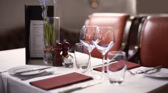 Elegant dukning i en restaurang med röda vadderade stolar. På bordet finns röda servetter, vinglas, bestick, en meny, salt- och pepparskakar och en vas med lila blommor, allt arrangerat på en vit duk.