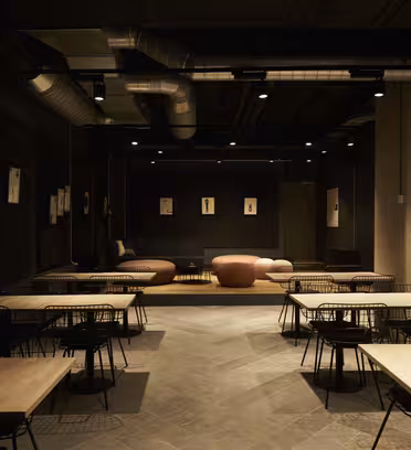 Ett svagt upplyst modernt café med svarta väggar och industriell inredning. Träbord och svarta trådstolar är snyggt ordnade. Bruna soffor finns på baksidan och små konstverk med ankartema hänger på väggarna. Taket har synliga kanalsystem.
