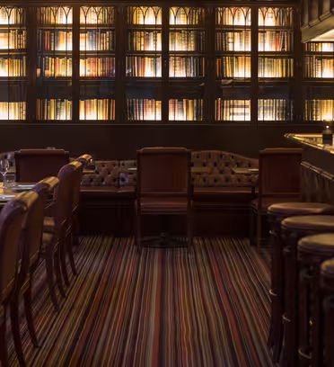 Mysig restaurang med bibliotekstema med svag belysning. Hyllor fyllda med böcker kantar väggarna. Läderstolar och en bar med pallar fyller utrymmet. Borden dukas med vinglas och fat på ett randigt heltäckningsmatta golv.