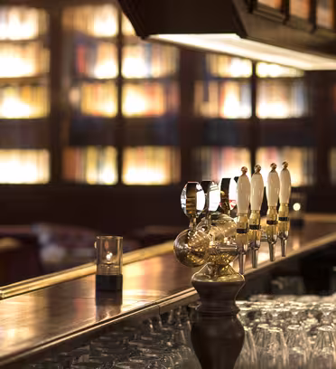 En mysig, svagt upplyst bar med en bänkskiva i trä, med flera ölkranar. Glasen ställs upp och ner på ett ställ nedanför. Hyllor fyllda med böcker är mjukt upplysta i bakgrunden, vilket skapar en varm och inbjudande atmosfär.