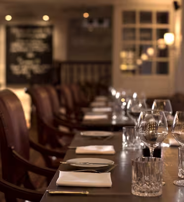 Svagt upplyst restauranginteriör med ett långt, elegant bord dukat för middagar. Läderstolar kantar bordet, som är snyggt arrangerat med vita servetter, kristallglas och bestick. En meny på den svarta tavlan är synlig i bakgrunden.