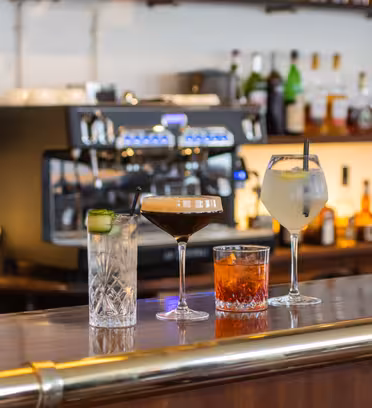 En barscen med fyra cocktails på disken: en gin och tonic med gurka, en espresso martini, en negroni och en vitvinssprit. I bakgrunden finns en kaffemaskin och olika spritflaskor på hyllorna.