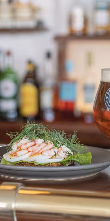 En tallrik med räkor och dill på bänk, tillsammans med en gaffel och ett glas bärnstensfärgat öl. Suddig bakgrund med en person nära en bar fylld med olika flaskor.