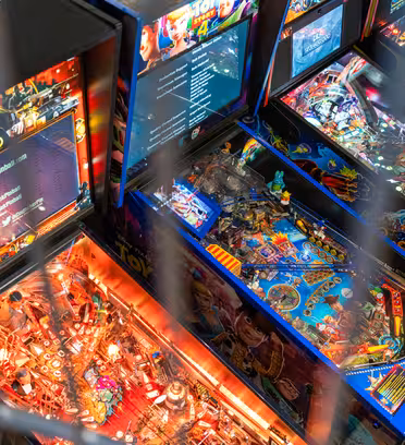 Fyra färgglada flipperspel med ljusa skärmar och intrikat design står uppradade sida vid sida. Glasskivorna avslöjar detaljerade spelfält med simfötter, stötfångare och olika temadekorationer. En metallstång skymmer delvis sikten.