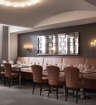 Elegant inredd matsal med bruna läderstolar och bord med vinglas. Rummet har en modern ljusarmatur, en stor spegel på väggen och en mjuk, neutral färgpalett.