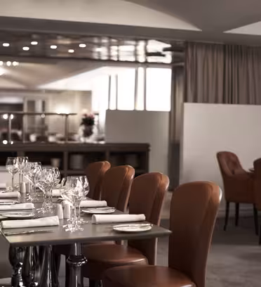 En modern, elegant restauranginredning med snyggt dukade bord. Bruna läderstolar omger bord prydda med glas, bestick och servetter. Mjuk belysning skapar en varm atmosfär och ett barområde syns i bakgrunden.