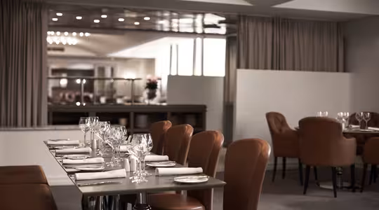 En modern, elegant restauranginredning med snyggt dukade bord. Bruna läderstolar omger bord prydda med glas, bestick och servetter. Mjuk belysning skapar en varm atmosfär och ett barområde syns i bakgrunden.