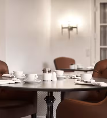 En mysig cafémiljö med ett runt bord omgivet av bruna läderstolar. Bordet är dukat med vita koppar, fat, servetter och en liten hållare som innehåller sötningsmedel. Bakgrunden har mjuk belysning och ett fönster.