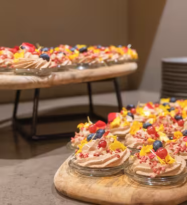 En visning av små desserter arrangerade på träfat. Varje dessert är prydd med blåbär, röda vinbär och dekorativa blommor. Högar av tallrikar syns i bakgrunden.