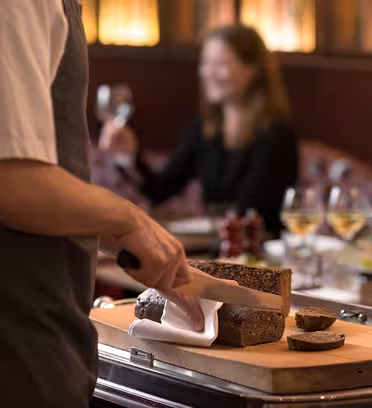 En person i ett förkläde skivar färskt bröd på en träskiva i en restaurang. En sittande individ i bakgrunden håller ett vinglas och det finns tre fyllda vinglas på bordet. Scenen är varm och elegant.