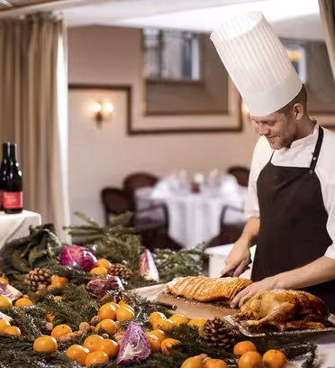 En kock i vit hatt och brunt förkläde snidar kött på ett bord som pryds av tallgrenar, apelsiner, rödkål och tallkottar. Vinflaskor finns i närheten. Inramningen är en varmt upplyst matsal med inramade konstverk på väggarna.