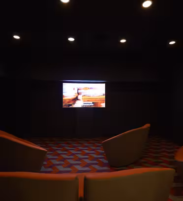 Ett svagt upplyst rum har en TV-skärm som visar en bild. Det står fyra moderna, orangea stolar mot skärmen på en mönstrad matta med röda och gråa geometriska former. Rummet har en mysig, minimalistisk atmosfär.