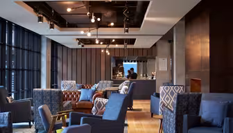 Modern lounge med blå och bruna stolar, en brun skinnsoffa och geometriskt mönstrade kuddar. Taklampor hänger ovanför, och en person arbetar bakom en bardisk. Stora fönster längs ena sidan släpper in naturligt ljus.