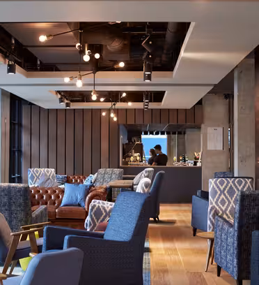 Modern lounge med blå och bruna stolar, en brun skinnsoffa och geometriskt mönstrade kuddar. Taklampor hänger ovanför, och en person arbetar bakom en bardisk. Stora fönster längs ena sidan släpper in naturligt ljus.