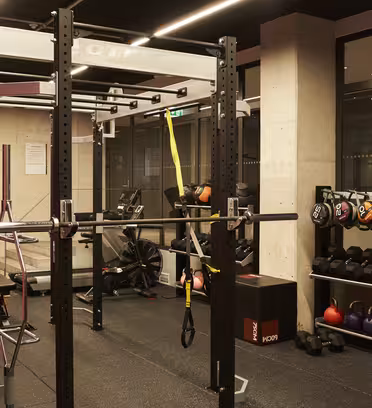 Ett modernt gym med ett squatställ med skivstång, motståndsband och upphängda remmar. Det finns kettlebells och hantlar snyggt organiserade på hyllorna, en motionscykel i bakgrunden och gummigolv överallt.