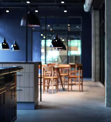 Modernt industrikök med en stor ö i förgrunden, med mörka skåp och en reflekterande yta. I bakgrunden syns träbord och stolar, upplysta av svarta taklampor. Betongväggar och stora fönster bidrar till den eleganta designen.