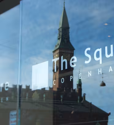 Reflektion av en historisk byggnad med en grön spira som ses i ett glasfönster. Orden "The Square" och "Copenhagen" syns på glaset. Himlen är blå med spridda moln.