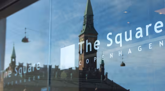 Reflektion av en historisk byggnad med en grön spira som ses i ett glasfönster. Orden "The Square" och "Copenhagen" syns på glaset. Himlen är blå med spridda moln.