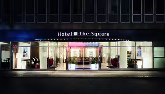 Nattvy av en hotellingång med modern design. På skylten ovan står "Hotel The Square". Glasdörrar och fönster avslöjar en elegant lobby med dekorativ belysning, konstverk och moderna möbler inuti.