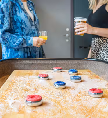 Två personer som står nära ett shuffleboardbord och håller drinkar. Bordet är sprinklat med puder och har röda och blå shuffleboard-puckar. Personen till vänster bär en blåmönstrad outfit, och den till höger bär en svart topp och en leopardmönstrad kjol.