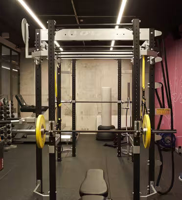Ett gym med ett squatställ, gula viktskivor, hantlar, kettlebells och bänkar. Det finns en rosa vägg med linjeteckningar av människor som tränar, och träningsutrustning är snyggt arrangerad. Utrymmet är väl upplyst med takbelysning.