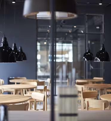Modern kaféinteriör med runda träbord och stolar. Stora svarta pendellampor hänger i taket. Neutrala färger och minimalistisk inredning skapar en mysig atmosfär. Glasväggar och betongdetaljer kompletterar utseendet.