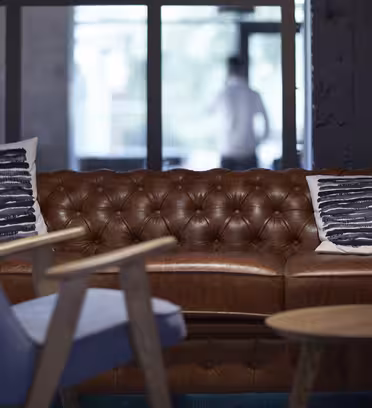 En mysig loungedel med en brun lädertuftad soffa med två mönstrade kuddar. I förgrunden finns en blå fåtölj och ett litet träbord. En suddig figur syns genom glasdörrarna i bakgrunden.