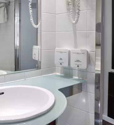 Modernt badrum med vitt handfat på glasbänkskiva, kromkran, väggmonterad hårtork och tvålautomater. Kaklade väggar och en handdukshylla syns. En spegel reflekterar en del av rummet, inklusive ett duschområde.