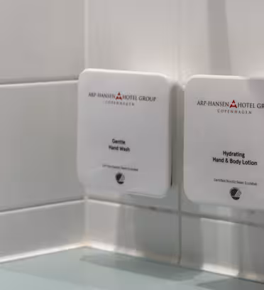 Väggmonterade dispensrar i ett badrum; en märkt "Gentle Hand Wash" och den andra "Hydrating Hand & Body Lotion." Båda är märkta med "ARP-HANSEN HOTEL GROUP COPENHAGEN." Reflexer synliga i den kaklade väggspegeln.