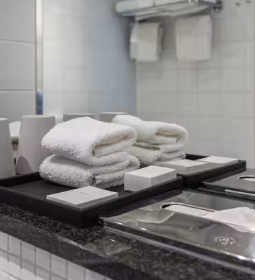 En bänkskiva i badrummet med snyggt arrangerade vita handdukar, två grå koppar och olika toalettartiklar. Under en spegel finns tissuelådor och en modern tvålautomat. Bakgrunden visar vikta handdukar på en hylla.