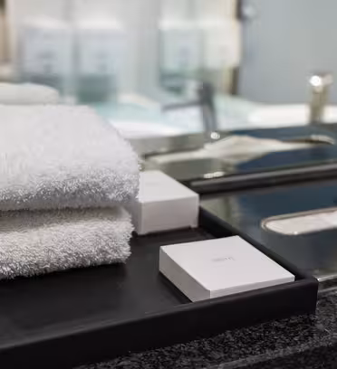 En prydligt arrangerad hög med vita handdukar på en svart bricka bredvid tvål, placerad på en mörk marmorbänk. En mjukpappershållare i blank metall och ett handfat med en spegel i bakgrunden kompletterar den eleganta badrumsmiljön.