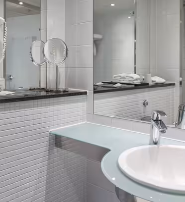 Modernt hotellbadrum med vitt kakel, en väggmonterad hårtork och en rund spegel. Bänkskivan har tvål och handdukar snyggt placerade. Diskbänken har en elegant kran, och det finns en glashylla ovanför en toalettpappershållare.
