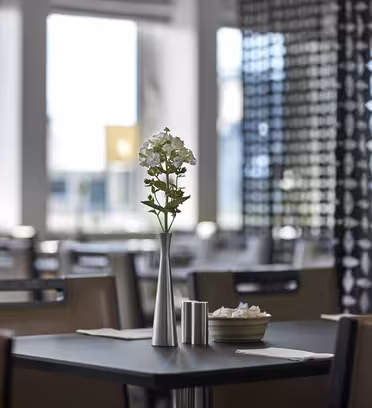 En modern caféinteriör med bord och stolar. En liten vas med vita blommor står på ett svart bord. Mjukt naturligt ljus filtrerar genom stora fönster och skapar en mysig och inbjudande atmosfär.