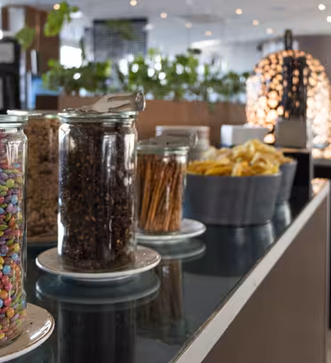 Burkar fyllda med färgglada godis, nötter och snacks kantar en buffédisk i ett modernt, väl upplyst rum. Bakom disken står växter och dekorativa ljusarmaturer, med stora fönster i bakgrunden.