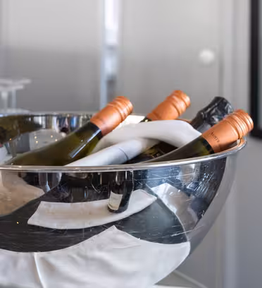 En ishink i silver rymmer flera flaskor vin, lutad för enkel åtkomst. Flaskorna har olika färgade halsar, främst orange. Vinglas är suddiga i bakgrunden och en delvis synlig vinflaska är i förgrunden.