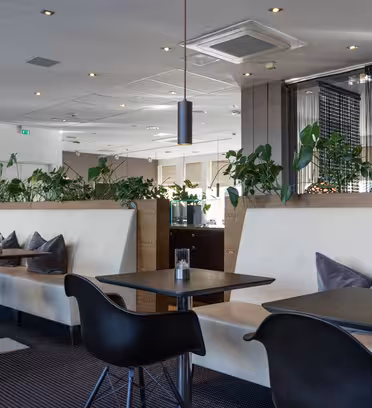 Modern caféinteriör med svarta bord och stolar, beige dämpade bås och gröna växter på skiljeväggar. Snygg belysning hänger i taket och skapar en mysig atmosfär. Stora fönster lyser upp utrymmet med naturligt ljus.
