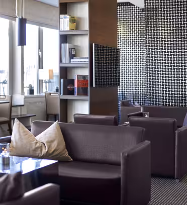 Modern lounge med mörkbruna soffor, låga bord och dekorativa kuddar. Hylla delar upp utrymmet, med böcker och inredning. Stora fönster till vänster ger naturligt ljus. Väggarna har geometriska mönster, vilket ger en modern känsla.