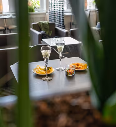 Två champagneglas och tallrikar med snacks, inklusive chips och nötter, sitter på ett bord i en mysig loungemiljö. Gröna växtblad ramar delvis in utsikten och mjuka fåtöljer syns i bakgrunden.