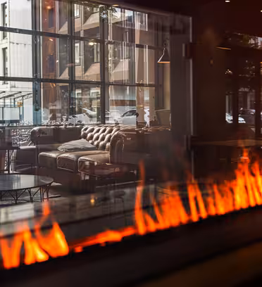 En modern lounge med skinnsoffor och fåtöljer, synligt genom ett stort glasfönster. I förgrunden skapar lågor från en elegant, horisontell eldstad en varm atmosfär. Reflexioner av stadsbyggnader syns i glaset.